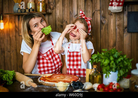 Happy mother and daughter wearing tabliers à carreaux pizza cuisine ensemble et s'amuser, faire face avec les yeux au lieu de tomates sur la table remplie d'ingrédients dans la cuisine en bois élégant Banque D'Images