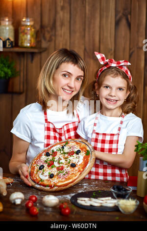 Happy happy mother and daughter wearing white T-Shirts et tabliers à carreaux montrant pizza cuite et looking at camera dans la belle cuisine en bois Banque D'Images