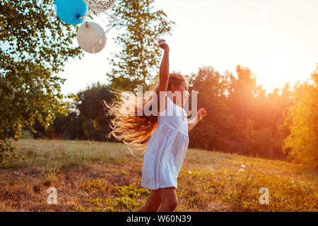 Happy little girl tournant avec baloons en main. Kid s'amuser dans le parc d'été au coucher du soleil. Banque D'Images