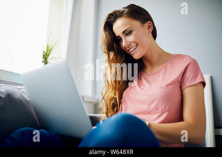 Portrait of a happy young woman smiling and using laptop while sitting on sofa in living room Banque D'Images
