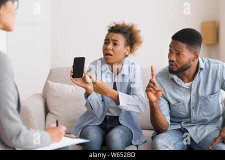 Femme Afro émotionnelle montrant Téléphone et blâmant mari pendant le traitement Banque D'Images