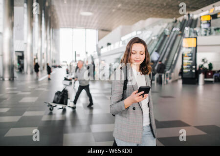 Jeune fille à l'aéroport promenades, regardant son smartphone en souriant. Banque D'Images