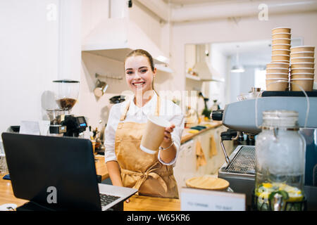 Young attractive woman smiling barista au comptoir bar, suggérant une tasse à café dans un café ou un restaurant moderne. Banque D'Images