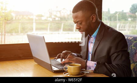 L'afro-américain d'affaires est en train de rédiger un message sur laptop sitting in cafe d'été près de la fenêtre. Homme noir écrit sur ordinateur, la lumière du soleil. Il porte en chemise et veston. Travailler avec tasse de café. Banque D'Images