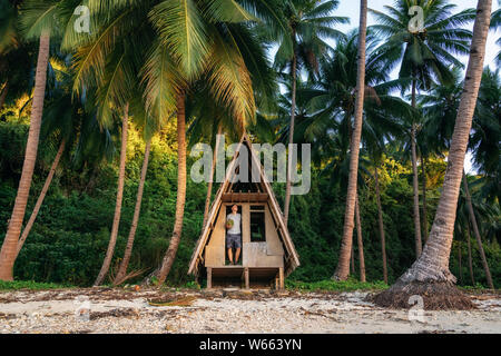 L'homme se tient à la porte de cabane et détient la noix de coco dans ses mains. La faune de la jungle à Port Barton, Philippines Banque D'Images