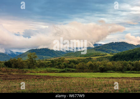 Domaine rural dans les montagnes au lever du soleil. arbres au bord d'un pré. temps couvert avec intéressant la formation de nuages au-dessus de la crête. début de l'automne Banque D'Images