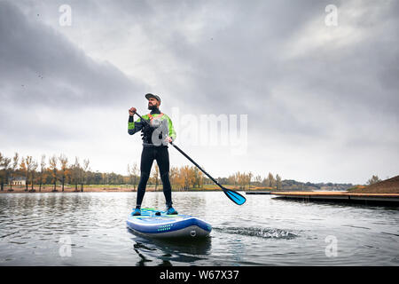 Athlète en combinaison sur paddleboard explorer le lac à froid contre ciel couvert