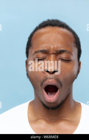 Close up portrait of young african-american man en chemise blanche sur fond bleu. Les émotions humaines, l'expression faciale, ad, ventes concept. Quelques secondes avant que le bâillement. Banque D'Images
