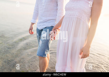 Tourné en plein air romantique de jeune couple en train de marcher le long de la côte se tenant la main. Jeune homme et femme marche sur la plage ensemble au coucher du soleil, corps clo