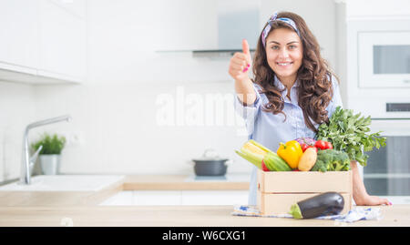 Femme dans sa cuisine avec boîte en bois plein de légumes organiques showing Thumbs up. Banque D'Images