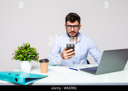 Young businessman in shirt se trouve en fonction à table et utilise le smartphone et boit du café sur le bureau avec ordinateur portable. Banque D'Images