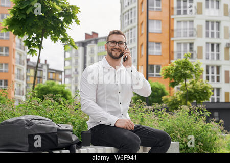 Jeune freelancer assis sur un banc, parler au téléphone, en regardant la caméra. Bel homme en portant des lunettes en blanc shirt élégant. Il worker smiling, posant sur fond de ville moderne. Banque D'Images