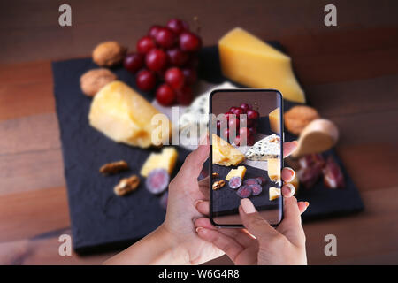 Mains de femme avec le smartphone taking photo assortiment de fromages, noix, raisins, fruits, viande fumée et un verre de vin sur une table de service. Et d'humeur sombre Banque D'Images