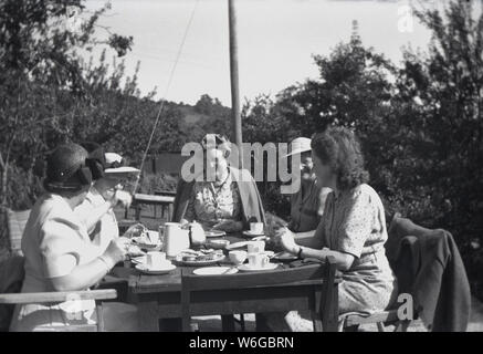 Années 1950, historiques, l'heure d'été et un groupe d'anglais habillé mesdames porter un chapeau s'amuser, prendre le thé et des scones et bonne coversation assis à une table en bois à l'extérieur, England, UK. Banque D'Images