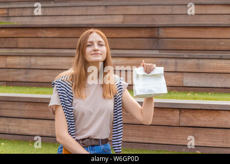 Smiling blonde student girl à t-shirt et jeans bleu est assis sur un banc en bois tenant un Doggy Bag sur une journée ensoleillée dans le parc. Pause déjeuner. Vide Banque D'Images