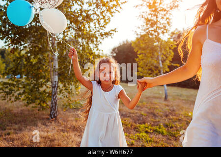 Heureux petite fille qui marche avec la mère et la tenue en main. baloons Family in parc d'été au coucher du soleil Banque D'Images