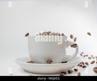 Café en grains tombent dans une tasse et soucoupe en porcelaine blanche, bondissant et sautant à travers l'air alerte transport l'excitation, toutes isolées contre un blanc Banque D'Images