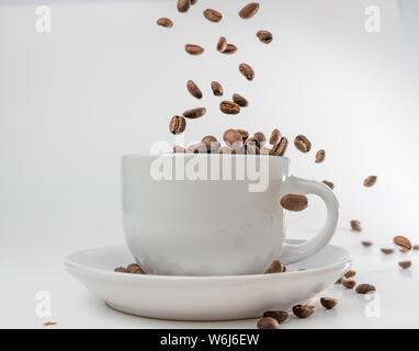 Café en grains tombent dans une tasse et soucoupe en porcelaine blanche, bondissant et sautant à travers l'air alerte transport l'excitation, toutes isolées contre un blanc Banque D'Images