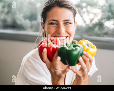 Caucasian girl sitting dans sa cuisine montrant trois gros poivrons de couleur. Vert, rouge et jaune. Banque D'Images
