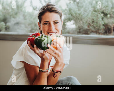 Caucasian girl sitting dans sa cuisine montrant trois gros poivrons de couleur. Vert, rouge et jaune. Banque D'Images
