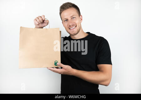 Beau jeune homme vêtu de noir T-shirt holding Shopping bag papper artisanat avec des marchandises isolées sur fond blanc Banque D'Images