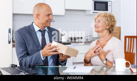 L'homme en costume d'affaires démontrant boîte en carton avec des marchandises commandées à mature woman in kitchen Banque D'Images