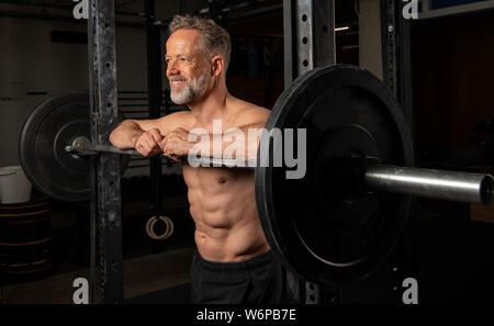 Portrait d'un athlète masculin plus attrayant avec des muscles forts. L'homme barbu souriant et athlétique est appuyé contre le rack d'haltères longs. Banque D'Images