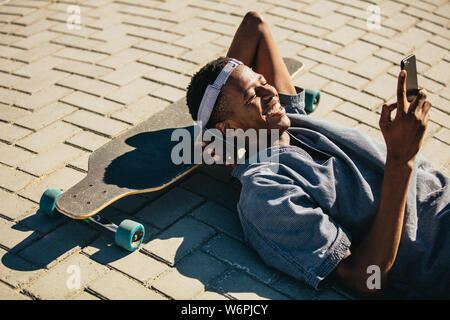 Smiling african american man allongé sur le skateboard et la lecture des messages texte sur son téléphone mobile. La planche à l'aide de smart phone tout en prenant un brea Banque D'Images