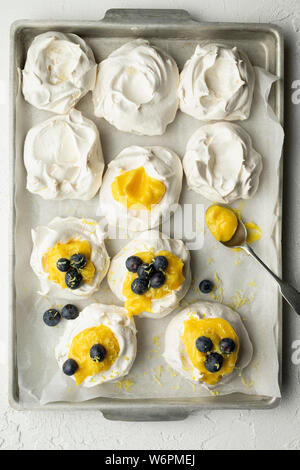 Des meringues avec le lait caillé de citron et bleuets sur une plaque à pâtisserie de papier sulfurisé, on a white background. Une cuillère de lait caillé de citron est sur le citron. Banque D'Images