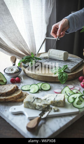 Un woman slicing fromage doux sur un plateau de fromages, avec divers légumes. Banque D'Images