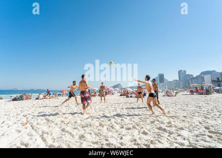 Groupe de jeunes jouant pied volley sur la plage de Copacabana sur une journée ensoleillée avec ciel bleu avec décor quartier typique Banque D'Images