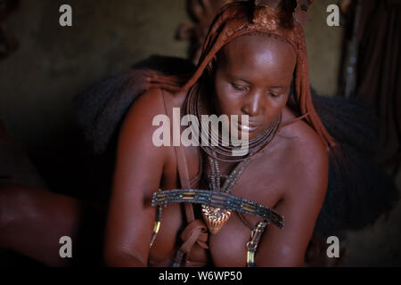 Opuwe, Namibie - Juillet 12, 2019 : une femme de la tribu Himba traditionnel l'accomplissement de sa beauté quotidien intérieur de kampor tree fumée et de poudre de pierre ocre ap Banque D'Images