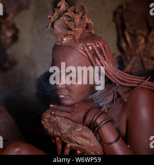 Opuwe, Namibie - Juillet 12, 2019 : une femme de la tribu Himba traditionnel l'accomplissement de sa beauté quotidienne la procédure de demande kampor tree fumer comme des douche repl Banque D'Images