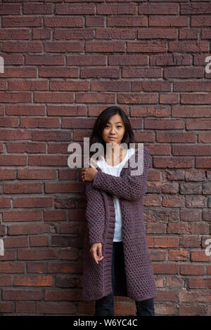 Belle asiatique Jeune femme debout dans un violet tricoté cardigan chaud, serrant elle-même sur l'épaule d'une main contre un mur permanent Banque D'Images