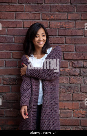 Smiling happy Asian girl in warm cardigan violet près de brick wall Banque D'Images