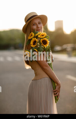 Belle jeune femme aux cheveux blonds en chapeau de paille sur la route en ville, tenant les mains bouquet de tournesols et looking at camera Banque D'Images