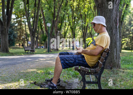 L'homme à chapeau et des lunettes à l'écoute de la musique s'asseoir sur un banc dans le parc. L'homme écoute de la musique avec le téléphone tout en vous relaxant dans le parc sous les arbres Banque D'Images