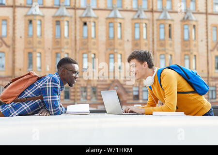 Vue latérale de portrait contemporain de deux étudiants de niveau collégial, de l'Afrique de l'un d'eux, souriant à l'autre debout sur les côtés opposés de table outdoors Banque D'Images
