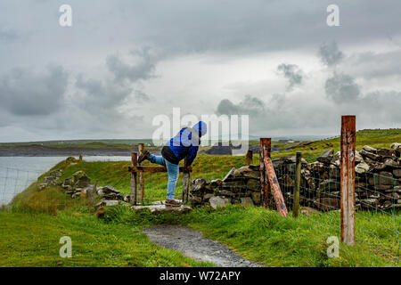 Femme sautant une clôture sur un jour de pluie sur la promenade côtière de Doolin route vers les falaises de Moher, et géosites geopark, façon sauvage de l'Atlantique, le printemps Banque D'Images