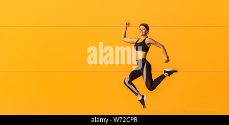 Vue latérale du jeune sportif attrayant satisfait à la mode sportswear femme en noir dans le mur orange. Mouvement de saut dynamique. Sp Banque D'Images