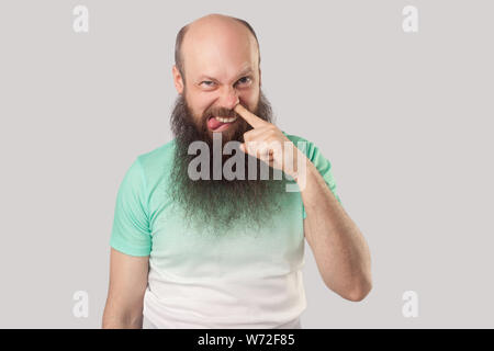 Portrait de drôle d'âge moyen sale homme chauve avec longue barbe en vert clair t-shirt forage permanent avec le doigt sur son nez, de la langue et à la recherche. Banque D'Images