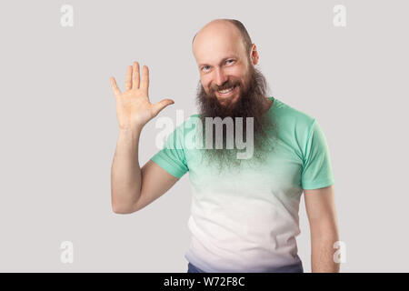 Portrait of happy senior homme chauve avec longue barbe en vert clair t-shirt standing looking avec sourire à pleines dents et en agitant sa main et d'accueil. ind Banque D'Images