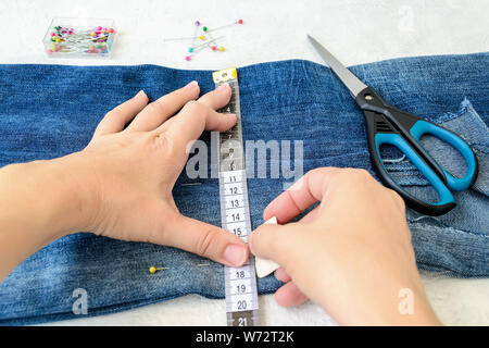 Femme mains dessinant une ligne de coupe sur un jean bleu. Jeans avec un grand trou sur une jambe du pantalon plié en deux et fixé avec les broches sur mesure. Raccourcir les jeans. D Banque D'Images