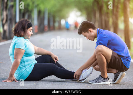 Blessures causées par le concept d'entraînement : asian man utiliser mains tiennent à sa cheville de grosse femme pendant le fonctionnement sur route dans le parc. L'accent sur la cheville. Banque D'Images