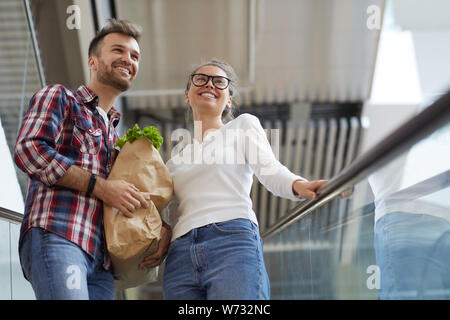 Low angle portrait of young couple riding escalator contemporain dans un centre commercial et un sac de transport, copy space Banque D'Images