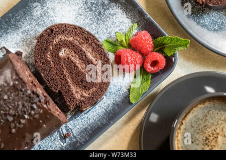 Swiss roll avec de la confiture au chocolat décoré avec des framboises dans une assiette et du café noir sur la table. Des bonbons. Focus sélectif. Banque D'Images