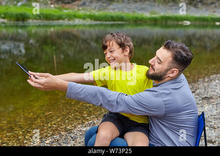 Jeune homme de race blanche barbu est assis près de la rivière avec son fils sur ses genoux et ils prennent des photos et souriant. Banque D'Images