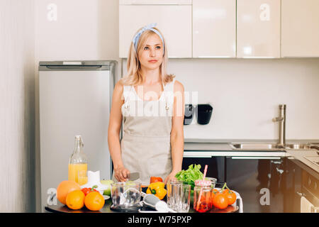 Young woman posing in kitchen, debout près de la table avec des fruits et légumes biologiques, souriant à la recherche d'appareil photo. Fille blonde wearing apron slicin Banque D'Images