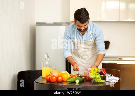 Les jeunes Indiens homme non rasé dans les verres, chemise bleue, tablier de la préparation des aliments sains et délicieux de légumes dans la cuisine le matin. L'équilibre Banque D'Images