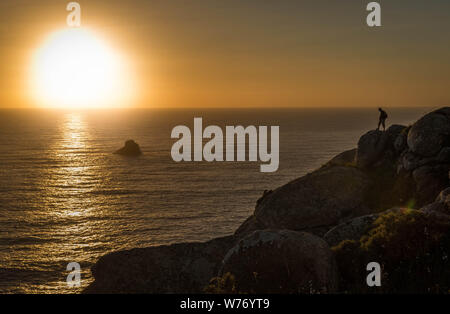 Pèlerinage sur le cap Finisterre, qui marque la fin du Camino de Santiago en Espagne. Silhouette d'un homme sur les rochers, face à la mer, et le coucher du soleil ligh Banque D'Images